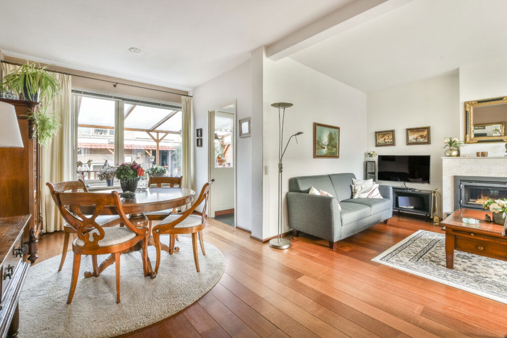 Bright living and dining room with wood floors, round table, sofa, TV, fireplace, and big window—pristine from expert San Mateo cleaning.